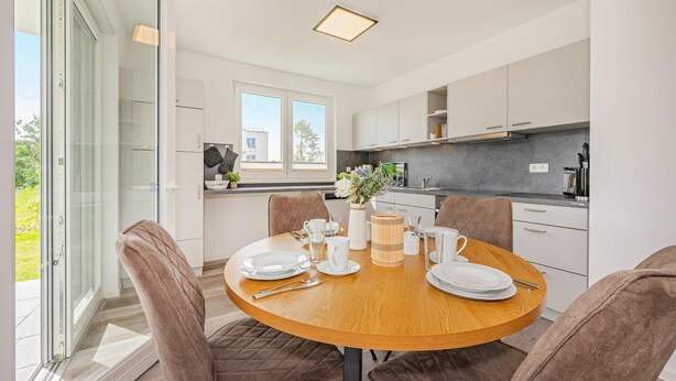 A modern kitchen with white cabinets, a round dining table set for four, and a large window providing a view of the outdoors.