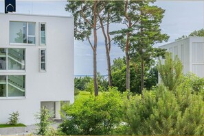Um edifício moderno com grandes janelas, uma escadaria visível e um jardim verdejante com árvores altas.
