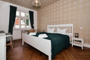A bedroom with a white bed, green bedding, a desk, a chair, a nightstand with a lamp, and a window with curtains. - Ferienwohnung Ole - Traumhafte Ferienwohnung mit Platz zum Arbeiten, Wlan und Terrasse (Heringsdorf)