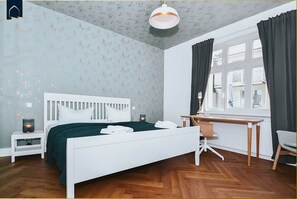 A bedroom with a white bed, a desk, a chair, a nightstand, and a window with curtains.