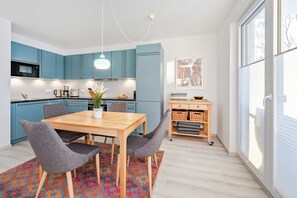 Une cuisine moderne avec des meubles bleus, une table à manger en bois avec des chaises, un luminaire suspendu et une vue dégagée grâce à de grandes fenêtres.
