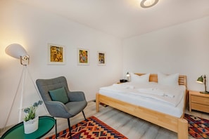 A bedroom with a bed, a chair, a bedside table, a lamp, and wall art.