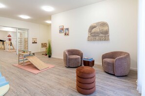 A room with a play area, two armchairs, a small table, and a wall-mounted tapestry.