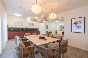 A modern dining area with a wooden table and wicker chairs, a kitchen in the background, and pendant lighting.