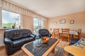 Un salon comprenant un canapé, des fauteuils, une table basse avec des fruits, et un coin repas avec une table et des chaises.