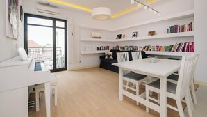 Et moderne, veloplyst rum med et klaver, et spisebord med stole, en reol og en sort sofa.