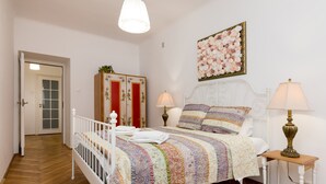 A bedroom with a white bed, a colorful quilt, two bedside lamps, a floral painting, and a wooden floor. - Royal Route Apartment for 10 people (Warszawa)