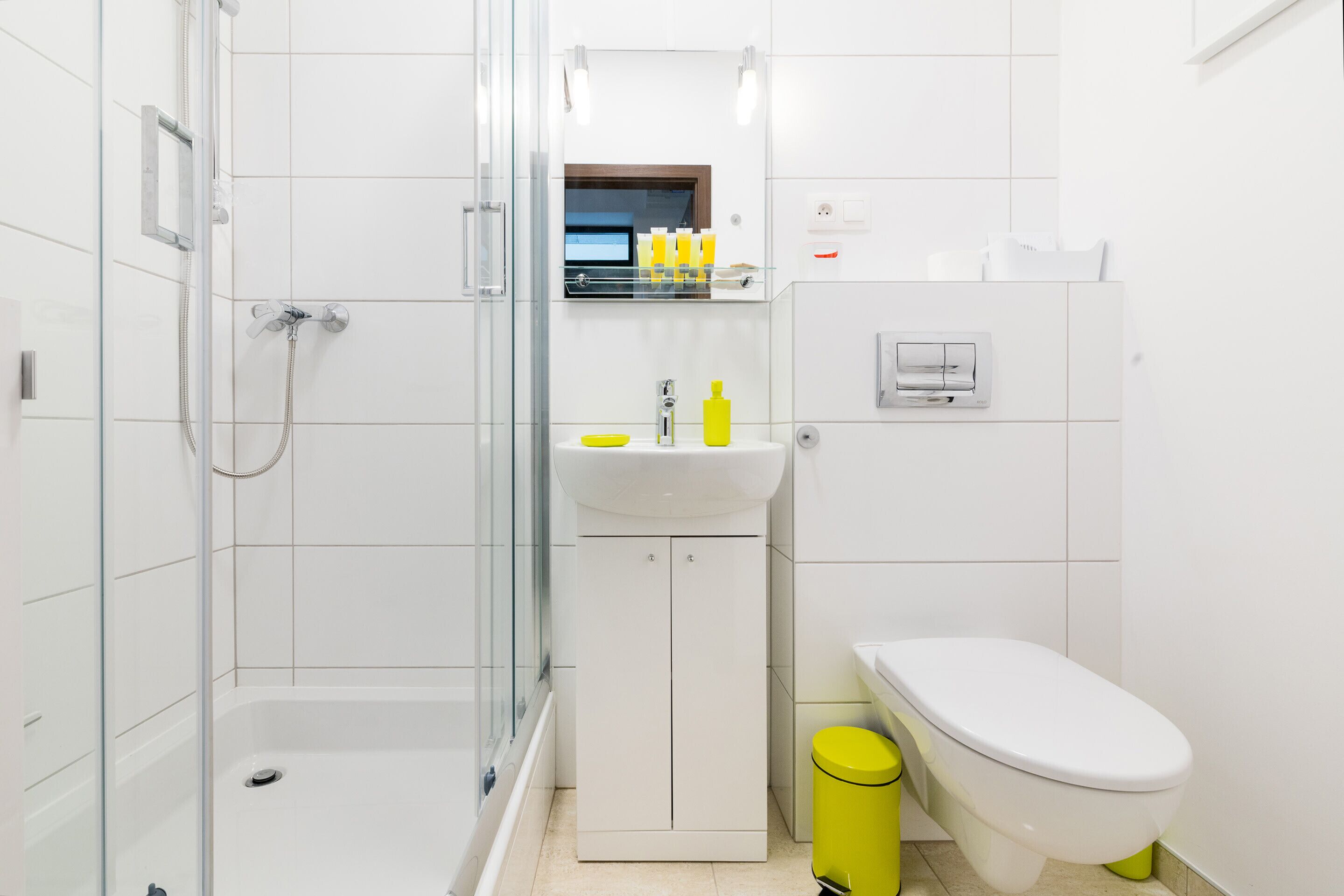 A modern bathroom with a glass shower enclosure, a white sink with a mirror above, and a toilet with a yellow trash bin.