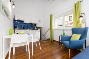 Un espace de vie compact comprenant un coin cuisine, un coin repas avec des chaises blanches, un fauteuil bleu et une fenêtre avec des rideaux verts.