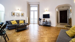 Una sala de estar con sofá, mesa de centro y sillas. Hay un televisor montado en la pared y un ventilador en el suelo.