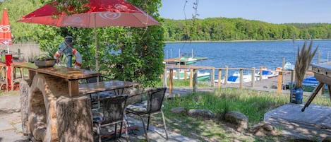 An outdoor seating area with a table and chairs, a red umbrella, and a view of a lake with boats docked.