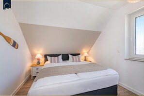 Quarto moderno com teto inclinado, uma cama grande com roupa de cama branca, duas mesas de cabeceira com abajures, e uma janela à direita.