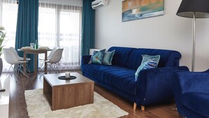 Uma sala de estar moderna com um sofá azul, uma mesa de centro de madeira e um tapete branco. Há uma área de jantar com mesa e cadeiras, e um quadro na parede.