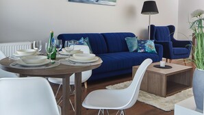 Uma sala de estar moderna com um sofá azul, uma mesa de jantar posta com pratos e copos, um quadro na parede e um abajur.
