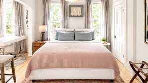 A bedroom with a large bed, a desk, and a chair. There are windows with curtains, a hanging light fixture, and a framed picture on the wall. - Historic Downtown-Old World Charm-Dining-Coffee (Delafield)