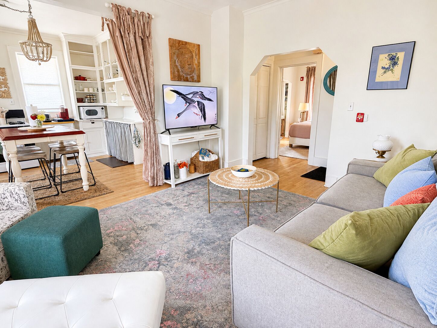 A living room with a grey sofa, a white armchair, and a coffee table. There is a television on a stand, a kitchen area with white cabinets, and a dining table with chairs.