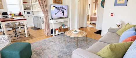Un salon avec un canapé gris, un fauteuil blanc et une table basse. On y trouve également un téléviseur posé sur un meuble, un coin cuisine avec des placards blancs, et une table à manger avec des chaises.
