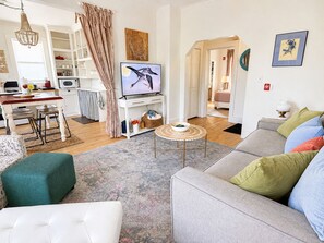 A living room with a grey sofa, a white armchair, and a coffee table. There is a television on a stand, a kitchen area with white cabinets, and a dining table with chairs. - Historic Downtown-Old World Charm-Dining-Coffee (Delafield)