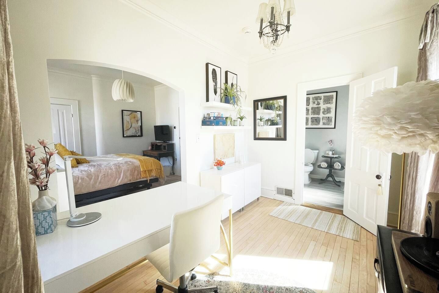 A spacious room with a large bed, a desk, and a chair. There is a door leading to another room with a toilet and a small table.