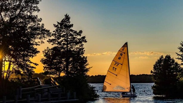 A sailboat with a sail marked with numbers on a body of water at sunset. - Historic Downtown-Old World Charm-Dining-Coffee (Delafield)