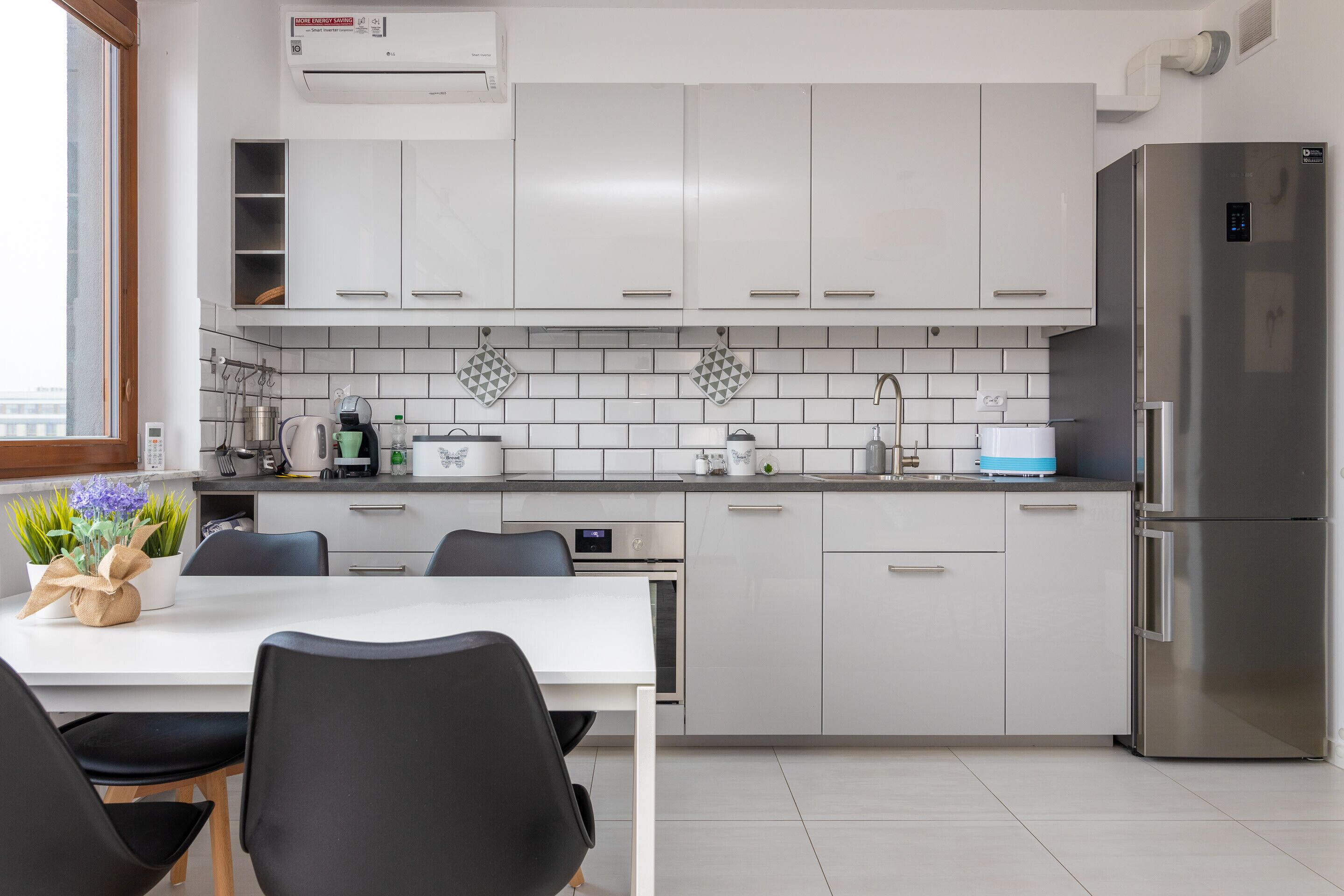 Una cocina moderna con armarios blancos, un refrigerador de acero inoxidable y un comedor con mesa y sillas.