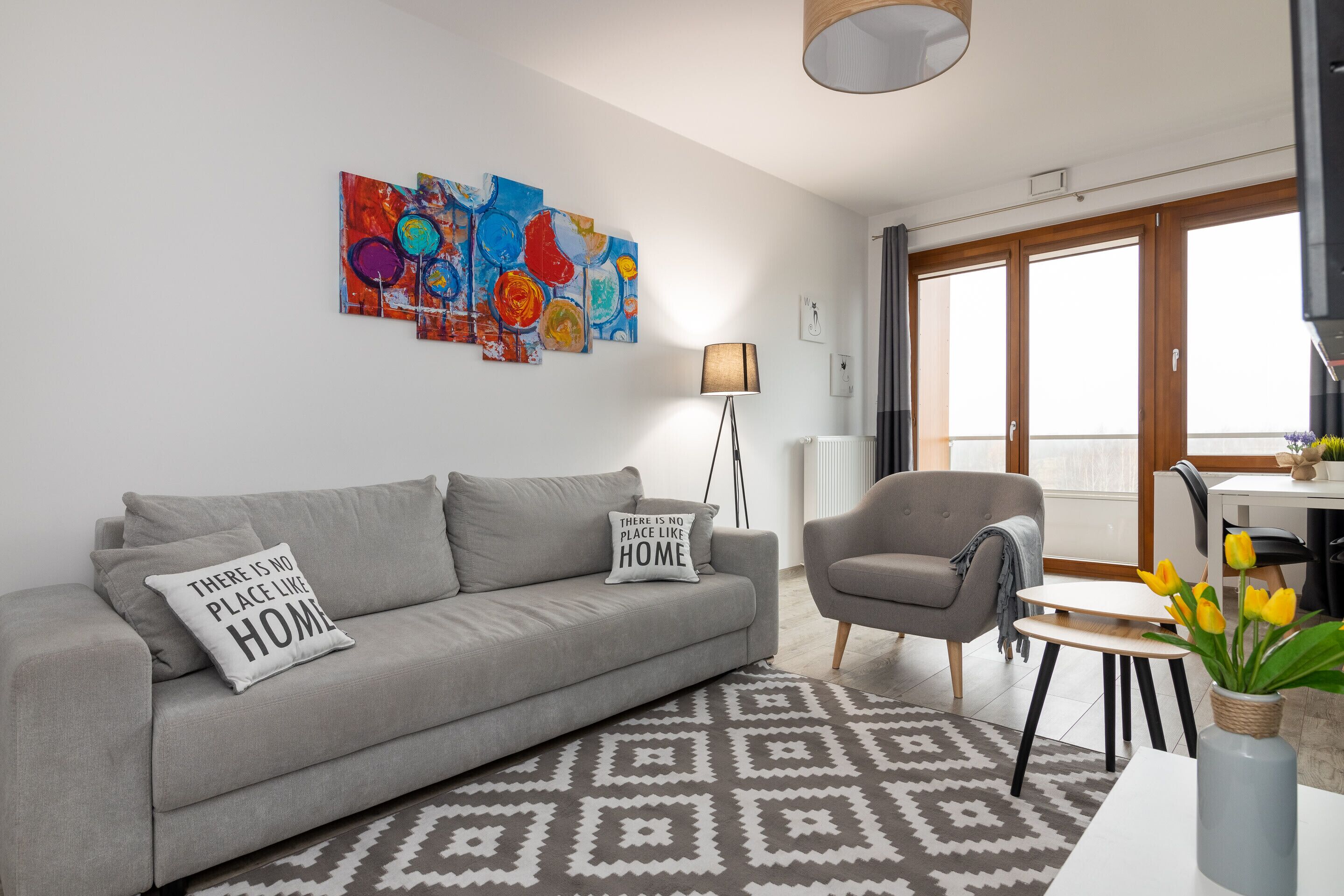 A modern living room with a grey sofa, a patterned rug, a grey armchair, and a table with a vase of flowers.