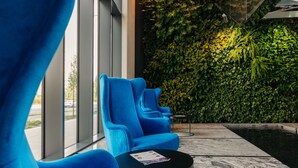 Un área de estar interior moderna con sillas azules y una pequeña mesa, una pared verde con plantas, y vistas al exterior a través de amplios ventanales.