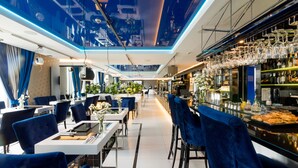 Un restaurante moderno con sillas de terciopelo azul, una zona de bar con copas de vino y un techo con diseño de paisaje urbano.