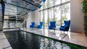 Una piscina interior con paredes de vidrio transparente, sillas azules y una escalera moderna.