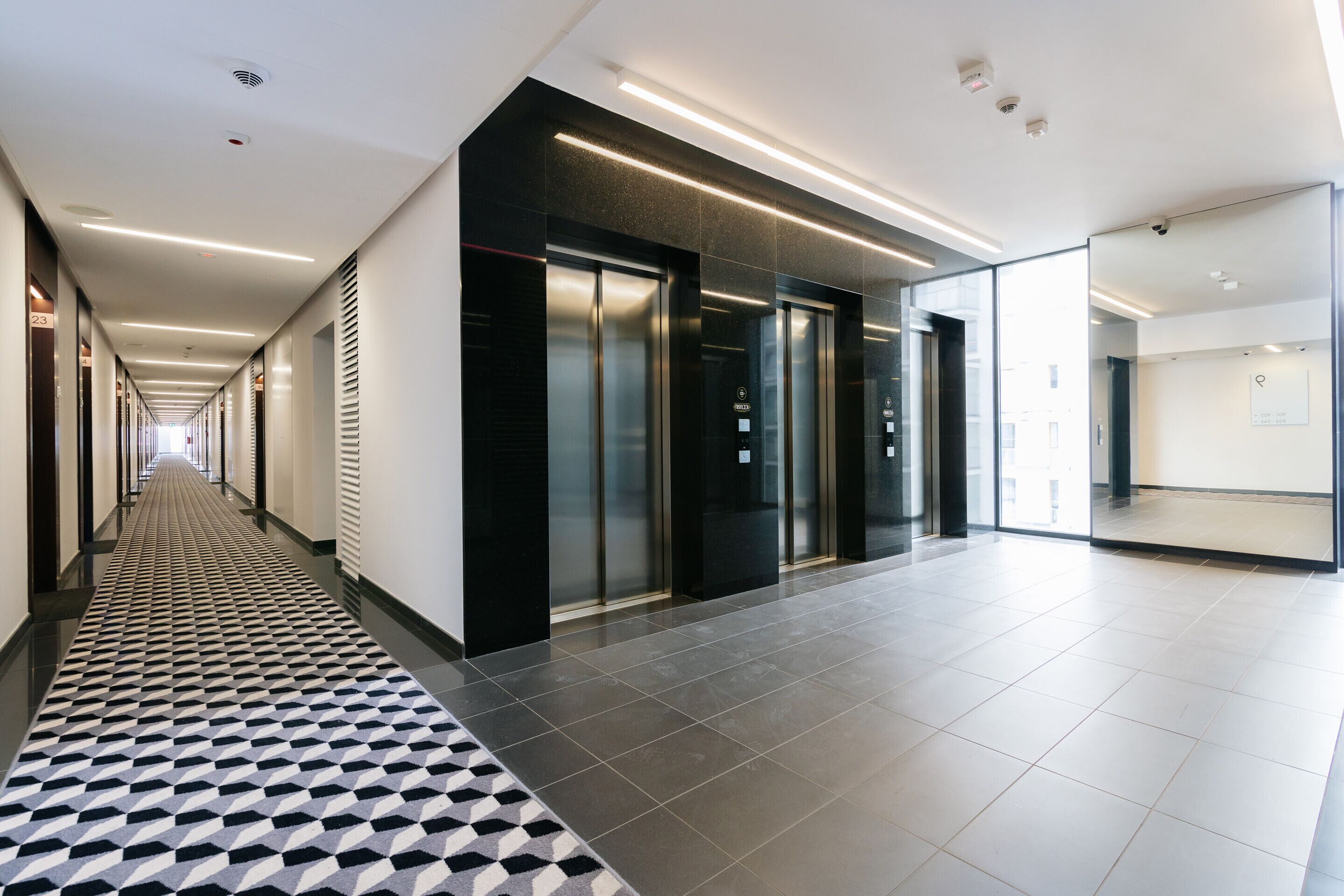 Pasillo moderno de hotel con alfombra estampada, ascensores y un muro de cristal.