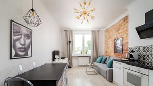 Een moderne keuken met een bakstenen muur, een eethoek met een zwarte tafel en transparante stoelen, en een zithoek met een grijze bank en een vloer met een patroon.