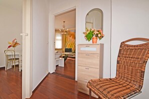 Ein Zimmer mit Holzboden, einer weißen Kommode mit Schubladen, einem Korbstuhl, einem Fernseher auf einem Standfuß und einem Spiegel an der Wand.