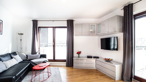 A modern living room with a black leather sofa, a red stool, and a flat-screen TV mounted on the wall.