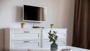 Ein modernes Wohnzimmer mit einem an der Wand befestigten Flachbildfernseher, einer weißen Kommode mit Schubladen, einer Glasvase mit Blumen auf einem weißen Tisch und einem braunen Vorhang im Hintergrund.