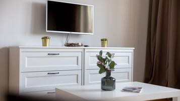 Un salon moderne avec un téléviseur à écran plat fixé au mur, un meuble blanc avec des tiroirs, un vase en verre contenant des fleurs sur une table blanche, et un rideau marron en arrière-plan.
