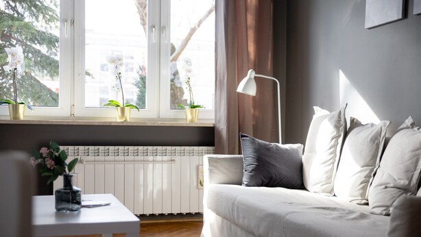 Ein modernes Wohnzimmer mit einem weißen Sofa, einem kleinen Tisch mit Blumenvase und einem Fenster mit zwei Blumentöpfen.