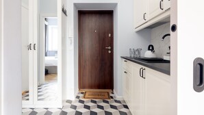 Een smalle gang met een houten deur, een deurmat en een keuken met witte kasten en een zwart aanrecht.