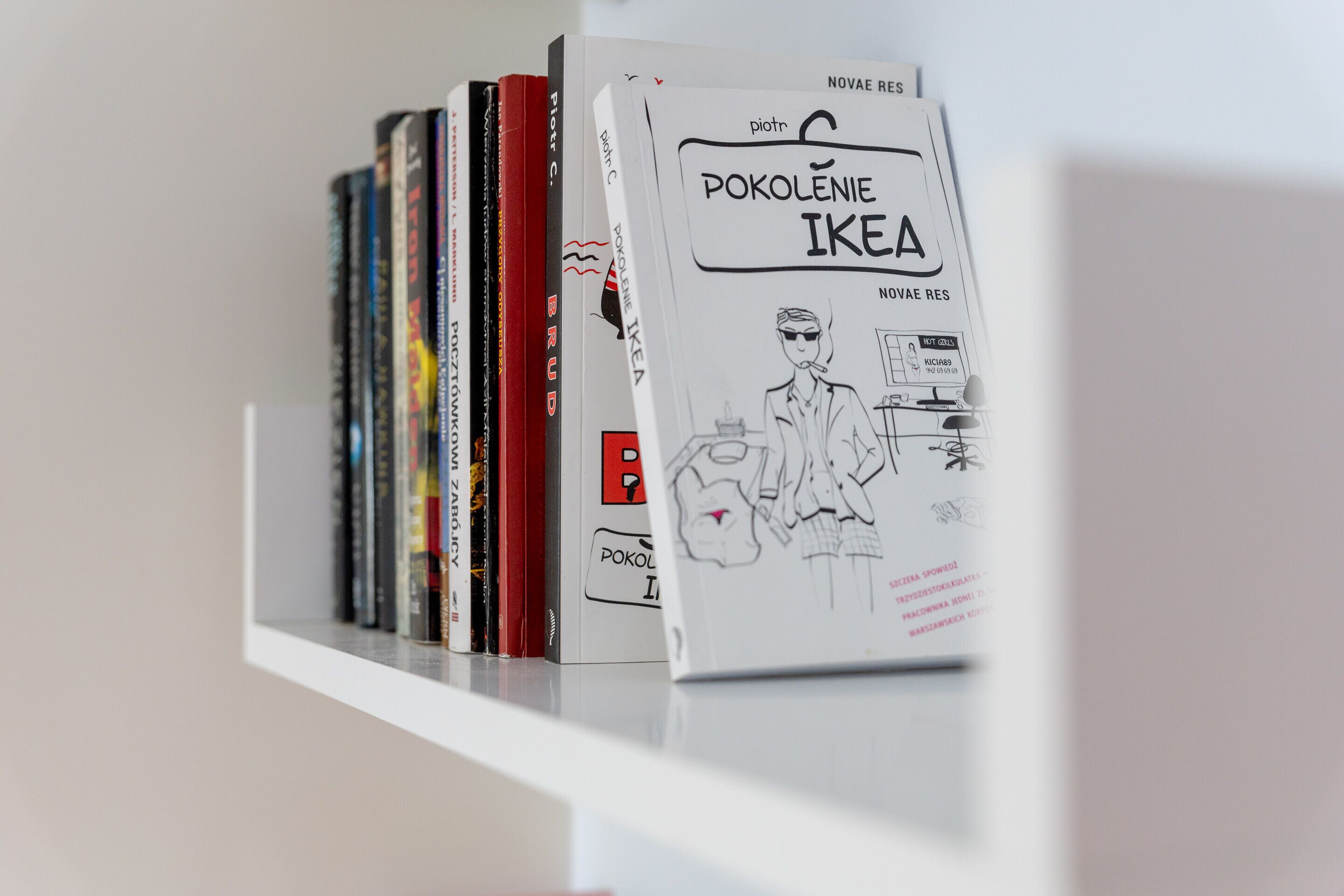 A bookshelf with various books, including a prominent book titled 'POKOLENIE IKEA' with a drawing of a person and IKEA products.