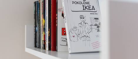 A bookshelf with various books, including a prominent book titled 'POKOLENIE IKEA' with a drawing of a person and IKEA products.