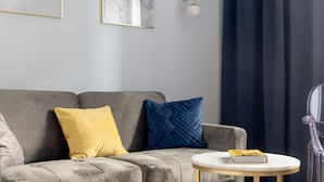 Een grijze bank met gele en blauwe kussens, een klein rond tafeltje met een boek, en een wandlamp.