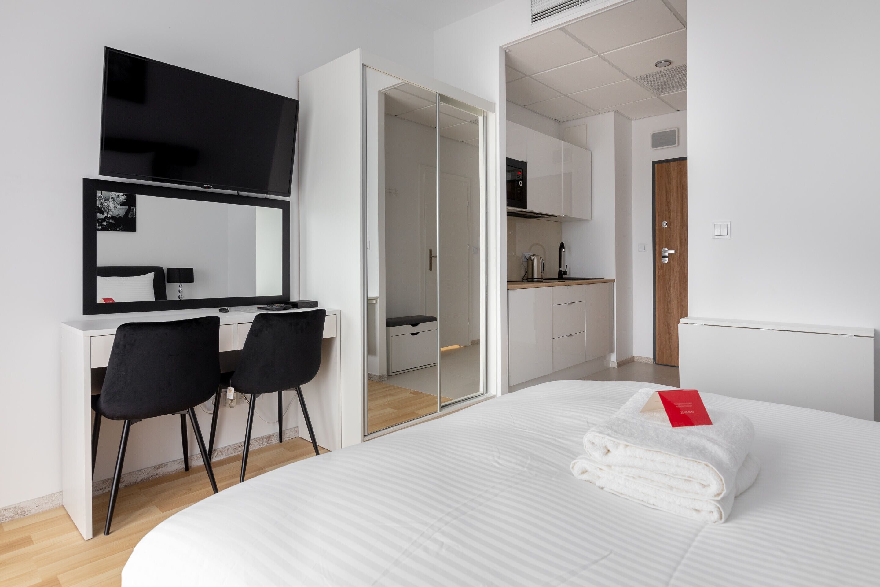 A modern hotel room with a bed, a desk with two chairs, a TV, and a sliding door leading to a kitchenette.