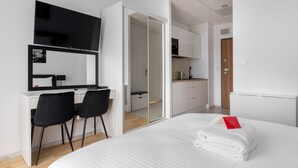 Moderni hotellihuone, jossa on sänky, työpöytä kahdella tuolilla, televisio ja liukuovi, joka johtaa pienelle keittiötilalle.