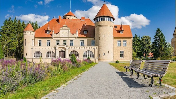 Un edificio que recuerda a un castillo, con una torre central, ventanas en arco y un camino empedrado que conduce a un banco.