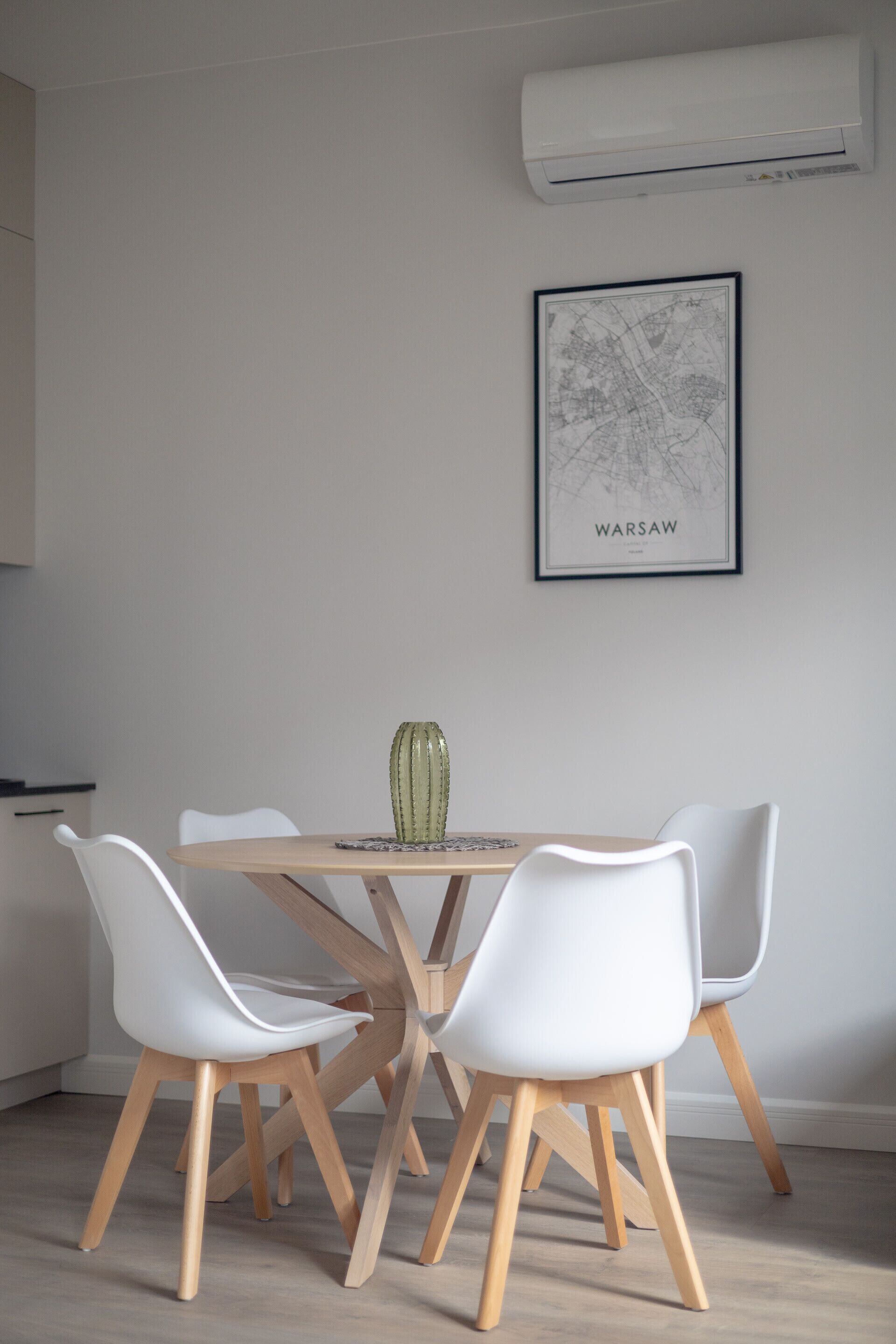 A dining area with a wooden table and white chairs, a green vase, and a framed map of Warsaw on the wall.