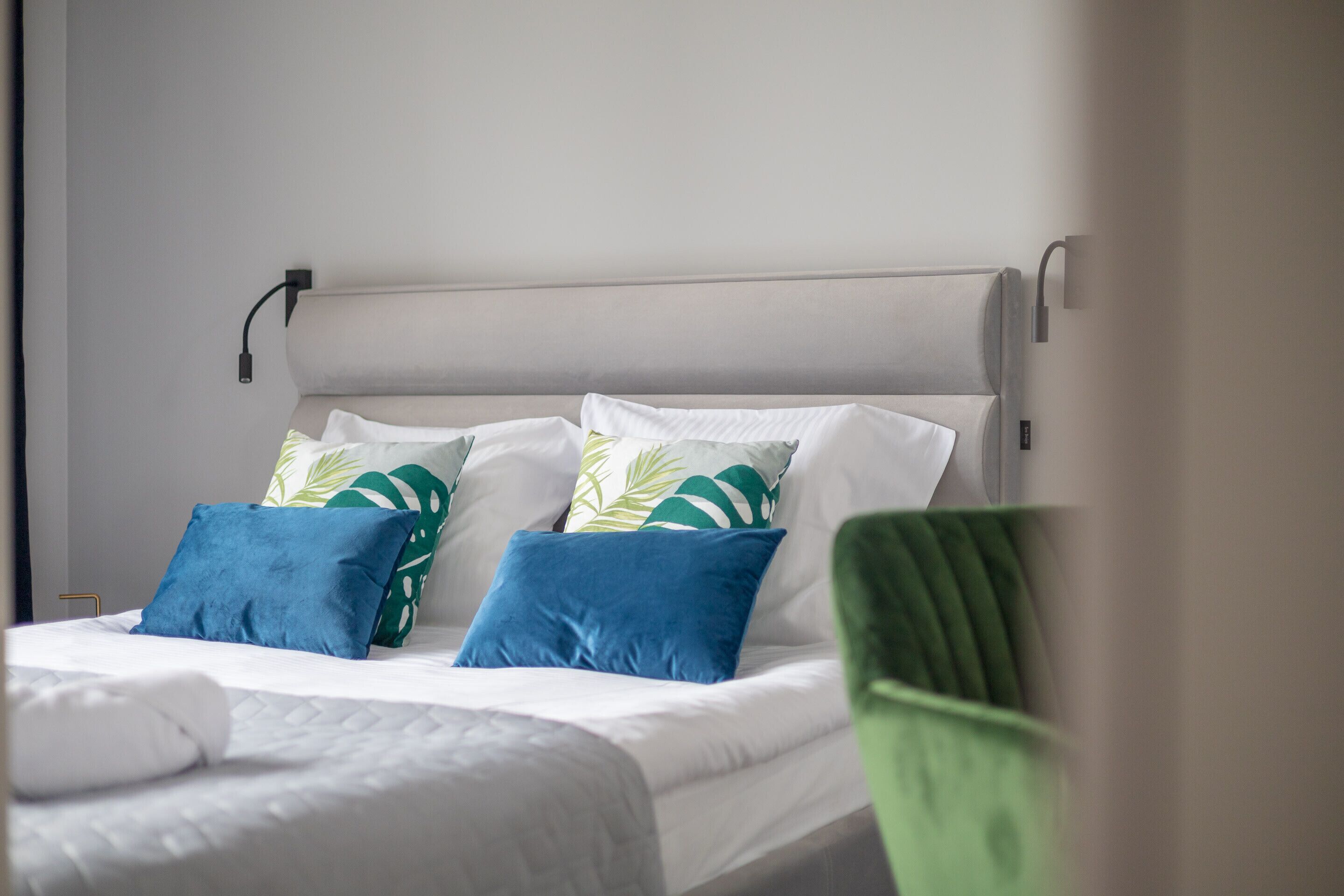 Un lit bien fait, avec du linge de lit blanc et des coussins aux motifs bleus et verts, une chaise verte et un luminaire fixé au mur.