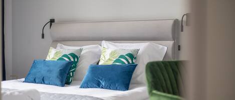 Un lit bien fait, avec du linge de lit blanc et des coussins aux motifs bleus et verts, une chaise verte et un luminaire fixé au mur.