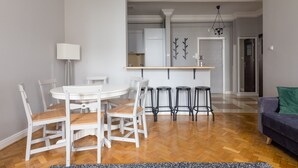 Een eetgedeelte met een witte ronde tafel en stoelen, een keuken op de achtergrond en een donkergrijze bank op de voorgrond.