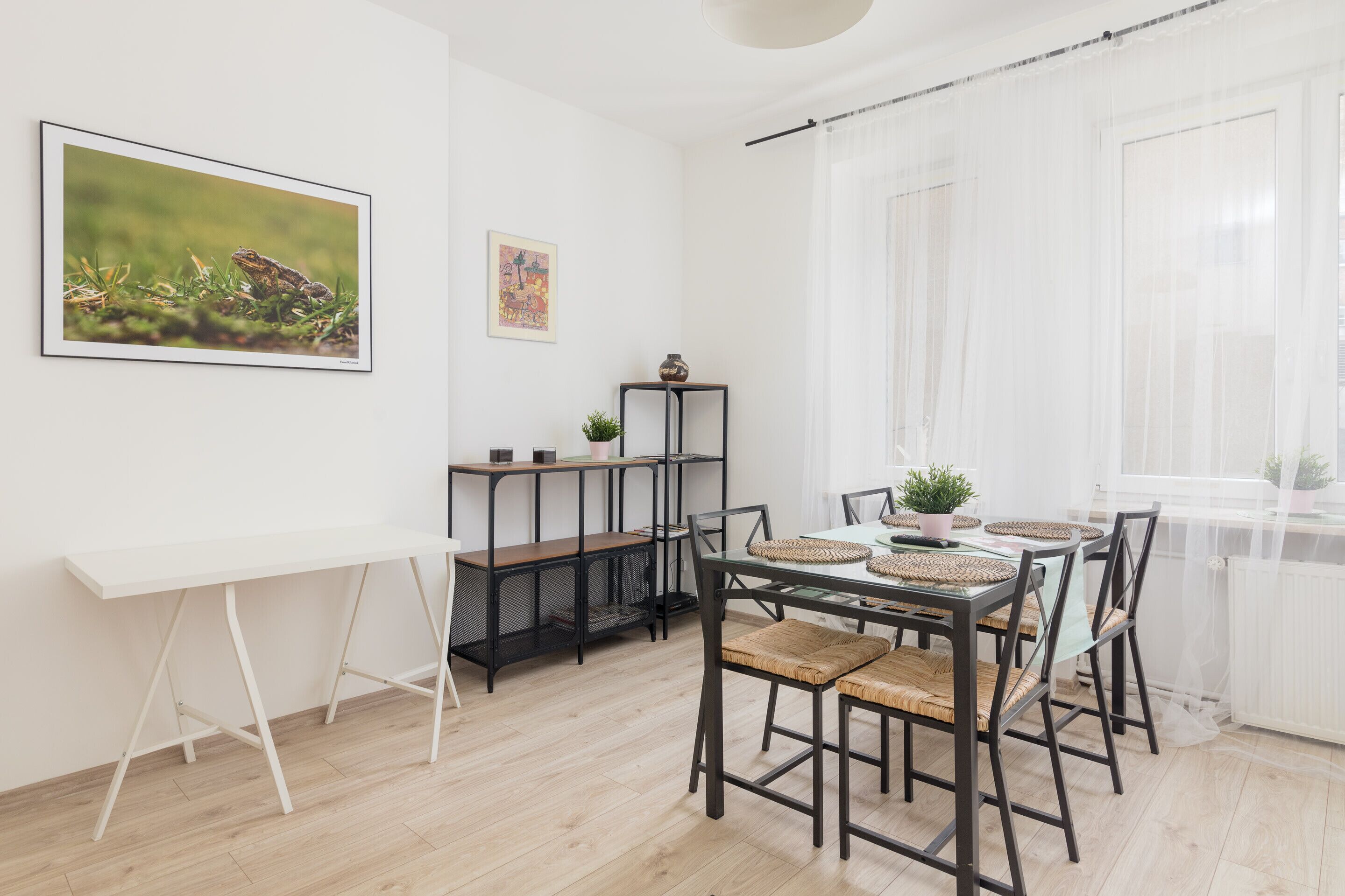 Een eetruimte met een witte tafel, zwarte stoelen en een whiteboard. Er is een raam met dunne gordijnen en een plant op de vensterbank.