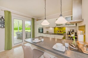 Une cuisine moderne dotée d’un grand îlot central, d’un coin repas avec des chaises et d’une vue sur un jardin grâce à des portes vitrées.