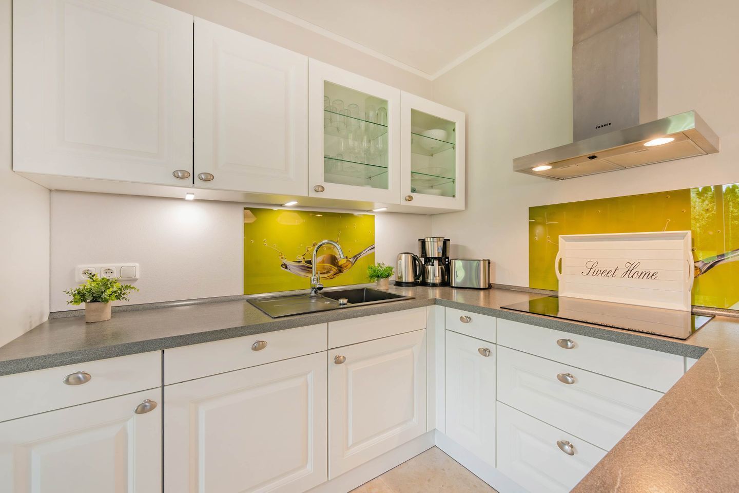 Una cocina moderna con muebles blancos, una campana extractora de acero inoxidable y una encimera con una cafetera y un letrero que dice 'Sweet Home'.